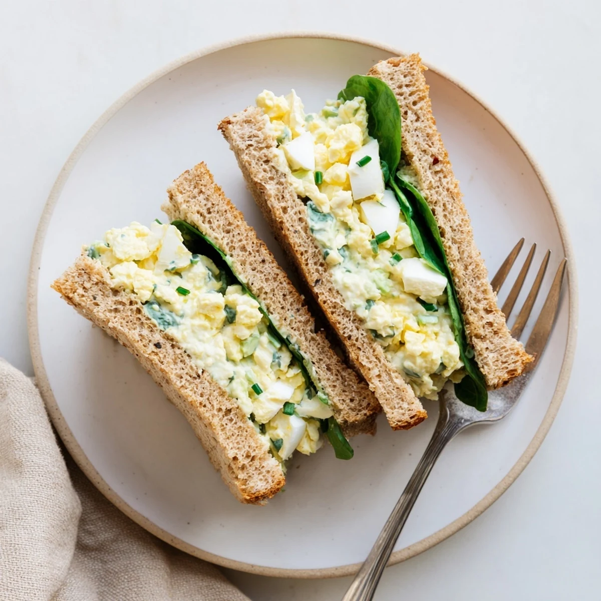 Freshly prepared Avocado Egg Salad Sandwich cut in half, revealing a hearty interior with spinach leaves and ripe tomato slices on the side.