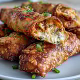 Freshly fried Buffalo Chicken Dip Egg Rolls rest on a cooling rack, steam rising from the crunchy, orange-tinted wrappers.
