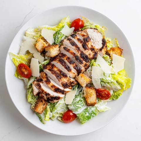 Tender grilled chicken strips fanning out over fresh chopped romaine, cherry tomatoes, and crunchy croutons in a salad bowl. A rich, creamy yogurt Caesar dressing is drizzled over the top, ready to toss.